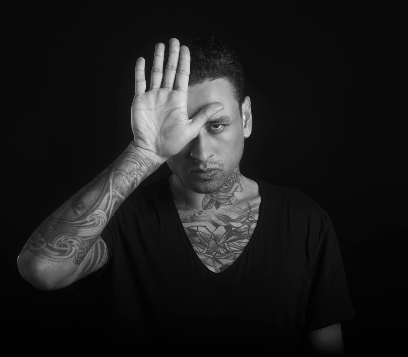 Black and white studio portrait of L7matrix against a dark ground, one open tattooed hand raised across half of his face.